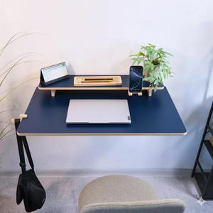 Peut inclure: Un bureau mural bleu marine avec une bordure en bois clair. Un ordinateur portable, un téléphone et une petite plante sont posés sur le bureau. Une petite étagère au-dessus contient un calendrier, des crayons et un plateau. Un sac noir est suspendu au bureau.