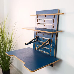 Könnte beinhalten: Ein blaues Wandbüro mit natürlichen Holzkanten. Es verfügt über eine Hauptarbeitsfläche, ein Regal und ein oberes Regal. Das Design umfasst Ausschnitte und abgewinkelte Stützen und bietet eine moderne, platzsparende Lösung.