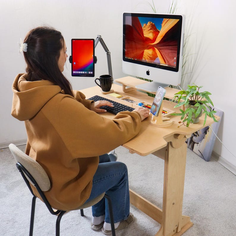 May include: A light-colored wooden standing desk with a computer, tablet, and phone. The desk has a keyboard, a small plant, and a black coffee mug. A person is sitting at the desk wearing a tan hoodie and blue jeans.