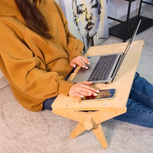 May include: A person sits on the floor using a wooden laptop stand with a laptop and a phone on it. The stand has a foldable design and is made of light wood.