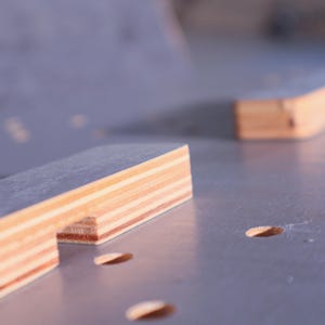 K&ouml;nnte beinhalten: Nahaufnahme von geschichteten Holzst&uuml;cken in hellbraunen und beigen Farbt&ouml;nen. Das rechteckige Holz ist Teil eines Bauprojekts. Der Hintergrund ist eine hellgraue Oberfl&auml;che mit kleinen, ovalen L&ouml;chern. Ma&szlig;e in Zentimetern.