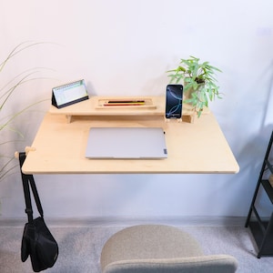 Op de afbeelding: Een lichtgekleurde, aan de muur gemonteerde bureau met een laptop, smartphone en kleine plant. Het bureau heeft een plank met potloden en een kalender. Een zwarte tas hangt aan het bureau en een grijze stoel staat eronder.