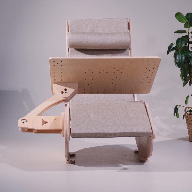 K&ouml;nnte beinhalten: Ein hellfarbiger ergonomischer Holzstuhl mit integriertem Schreibtisch. Der Stuhl verf&uuml;gt &uuml;ber eine gepolsterte Kopfst&uuml;tze, Sitzfl&auml;che und R&uuml;ckenlehne aus neutralem Stoff. Der Schreibtisch ist verstellbar und hat viele kleine L&ouml;cher. Der Stuhl ist auf Komfort und Funktionalit&auml;t ausgelegt.