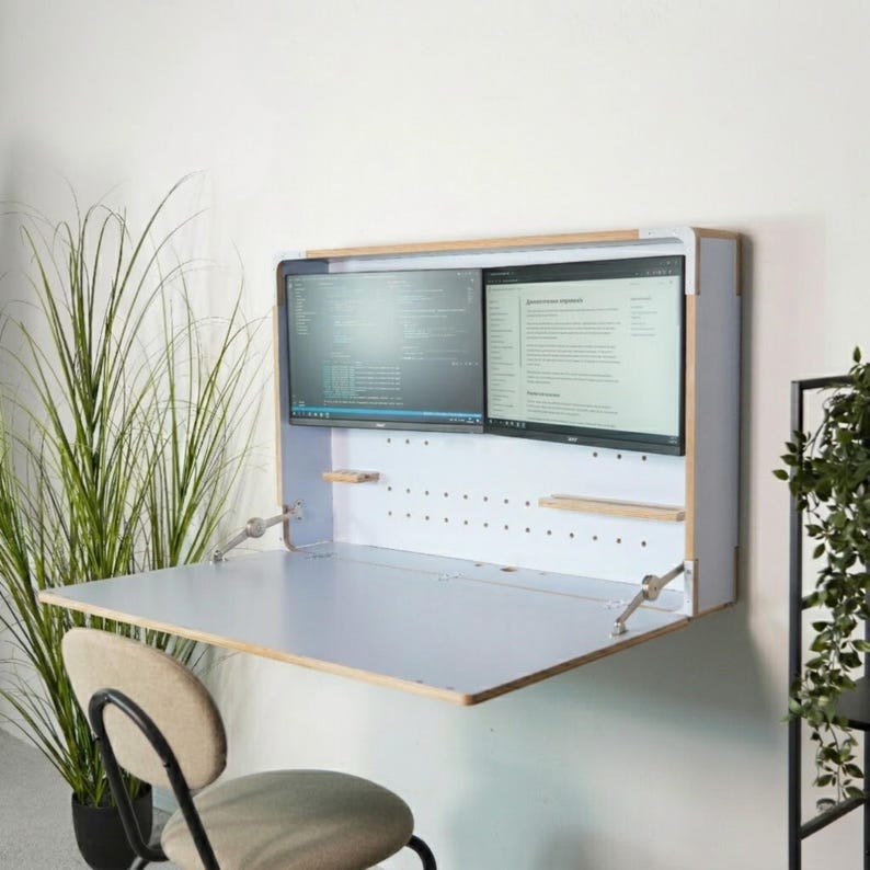 May include: A wall-mounted, fold-down desk featuring a light-colored wooden frame, a white pegboard back panel, and a flat work surface. Two computer monitors are mounted inside the unit, displaying text and code against a white wall.