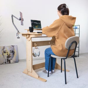 May include: A person sits at a light wood standing desk with a laptop, a monitor, and a cup of coffee. The desk has a shelf for storage and a hook for headphones. The person is wearing a brown hoodie and blue jeans.
