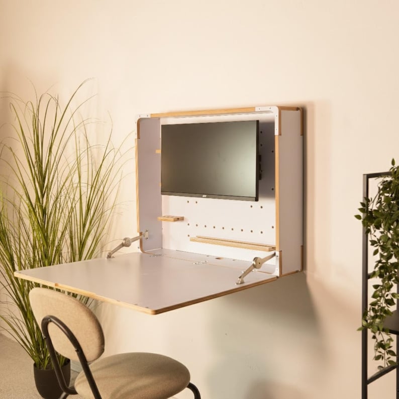 May include: A wall-mounted, fold-down desk unit featuring a light wood frame and white interior panels. The unit includes a built-in black computer monitor, small wooden shelves, and metal support hinges that hold the desk surface in a horizontal position.