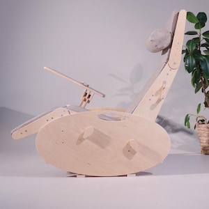 K&ouml;nnte beinhalten: Ein hellfarbiger Schaukelstuhl aus Holz mit eingebautem Schreibtisch und Kopfst&uuml;tze. Der Stuhl hat eine geschwungene Basis und eine verstellbare R&uuml;ckenlehne und Beinauflage. Die Kopfst&uuml;tze ist ein abgerundetes, gepolstertes Kissen.