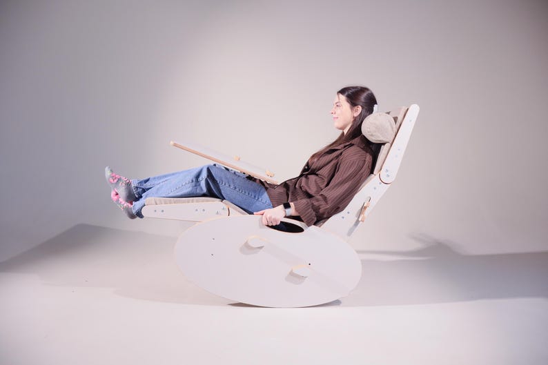May include: A light-colored wooden rocking chair with a headrest and a footrest. The chair has a unique design with a curved base and a wooden tray. The person is wearing blue jeans and a brown shirt.