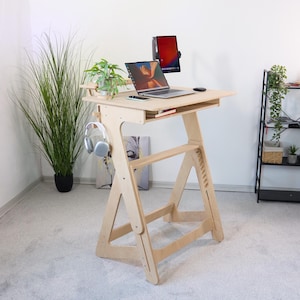 May include: A light-colored wooden standing desk with a laptop, tablet, and headphones. The desk has a shelf for plants and a drawer. The adjustable height desk is designed for ergonomic use and has a minimalist design.