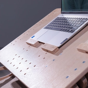 K&ouml;nnte beinhalten: Ein helles Laptop-Schreibtisch aus Holz mit einem Laptop und einem Tablet. Der Schreibtisch hat eine perforierte Oberfl&auml;che und Holzst&uuml;tzen f&uuml;r Laptop und Tablet. Der Laptop ist silber und schwarz.