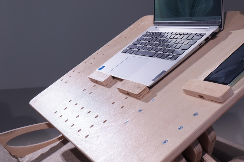K&ouml;nnte beinhalten: Ein helles Laptop-Schreibtisch aus Holz mit einem Laptop und einem Tablet. Der Schreibtisch hat eine perforierte Oberfl&auml;che und Holzst&uuml;tzen f&uuml;r Laptop und Tablet. Der Laptop ist silber und schwarz.