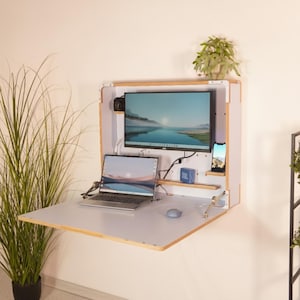 Könnte beinhalten: Ein weißer, an der Wand montierter, herunterklappbarer Schreibtisch mit Laptop, Monitor und Smartphone. Der Schreibtisch hat eine helle Holzverkleidung. Ein kleiner Lautsprecher und eine Pflanze sind ebenfalls sichtbar. Der Monitor zeigt ein Landschaftsbild.