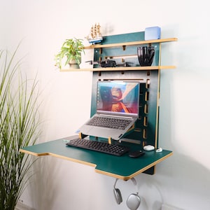 Op de afbeelding: Een aan de muur gemonteerd, donkergroen bureau met een laptop, toetsenbord en muis. Het bureau heeft twee planken met decoratieve items, waaronder een kleine plant, een speelgoedzeilboot en een zwarte mok. Een koptelefoon hangt eronder.