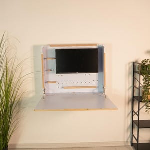 May include: A wall-mounted white desk with a fold-down surface and a light wood frame. The unit features a central black monitor screen, small wooden side shelves, and a perforated back panel. Metal hinges support the folding desk surface.