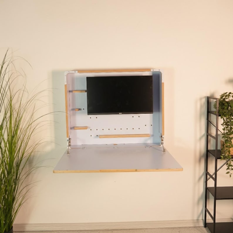 May include: A wall-mounted white desk with a fold-down surface and a light wood frame. The unit features a central black monitor screen, small wooden side shelves, and a perforated back panel. Metal hinges support the folding desk surface.