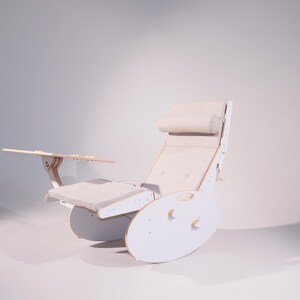 May include: A white wooden rocking chair with a built-in side table and a headrest. The chair has a light-colored cushion and a curved base. The side table is attached to the armrest and is made of wood.