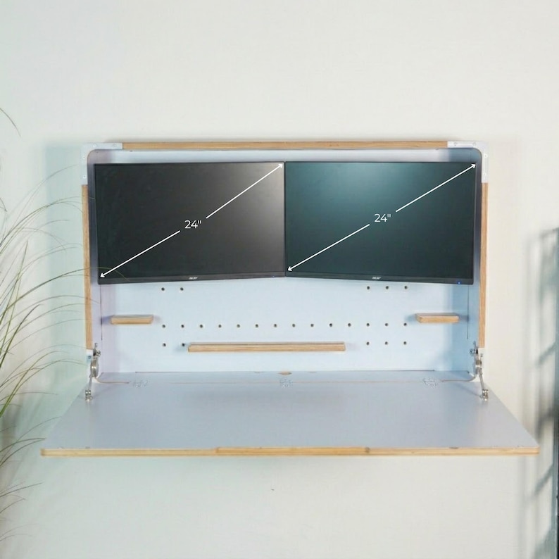 May include: A wall-mounted fold-down desk with a white back panel featuring pegboard holes and light wood trim. Two black 24-inch monitors are mounted inside the unit. The desk surface is white with light wood edges and metal hinges.