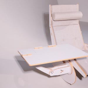 May include: A modern, light-colored lounge chair with an attached, adjustable white table. The chair has a padded headrest and seat, with a wooden frame. The table is rectangular and has a light wood trim.