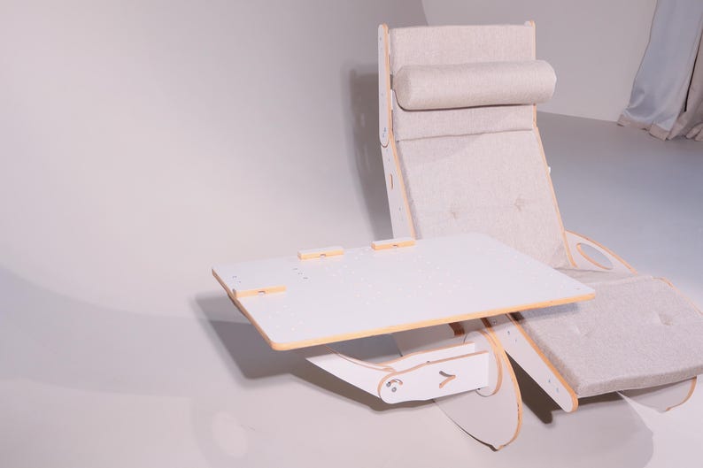 May include: A modern, light-colored lounge chair with an attached, adjustable white table. The chair has a padded headrest and seat, with a wooden frame. The table is rectangular and has a light wood trim.