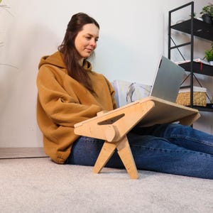 May include: A light-colored wooden laptop desk with folding legs. The desk is designed to be used while sitting on the floor. The laptop is silver and open. The person is wearing a brown hoodie and blue jeans.