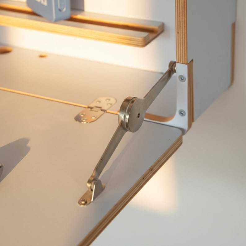 May include: A light gray drop-leaf desk surface featuring a metallic folding support hinge mechanism. The desk is constructed from plywood with visible layered edges, and a small metal hinge connects the flat surface to the main frame.