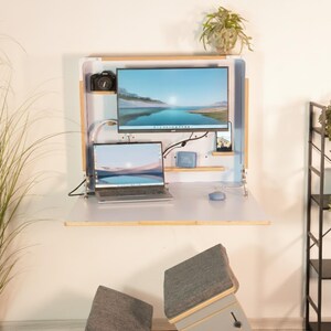 May include: A wall-mounted, fold-down wooden desk featuring a white interior panel, a mounted computer monitor, a laptop, a small camera, a blue speaker, and a smartphone. The desk includes integrated shelving and a light-colored wood frame.