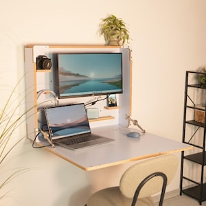 May include: A wall-mounted, fold-down desk with a monitor, laptop, and camera. The desk is white with a light wood trim and has a small shelf. A small plant sits on top of the desk unit. A gray chair is in front of the desk.