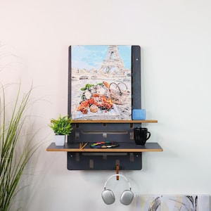 May include: A black wall shelf with two shelves, a painting of the Eiffel Tower, a small potted plant, a blue soap dish, a black mug, a set of paintbrushes, a palette of watercolors, and a pair of white headphones hanging from a hook.
