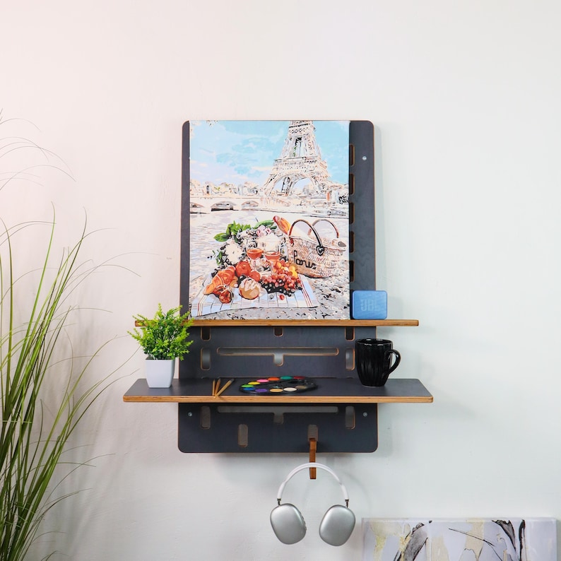 May include: A black wall shelf with two shelves, a painting of the Eiffel Tower, a small potted plant, a blue soap dish, a black mug, a set of paintbrushes, a palette of watercolors, and a pair of white headphones hanging from a hook.