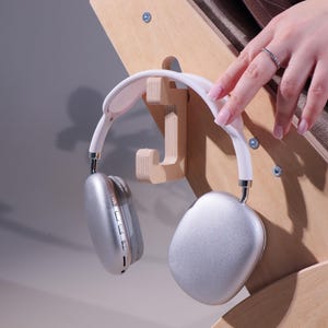 May include: Silver and white over-ear headphones hanging on a wooden headphone stand. The headphones have a white headband and silver earcups. The stand is made of light-colored wood and is mounted on a wooden surface.