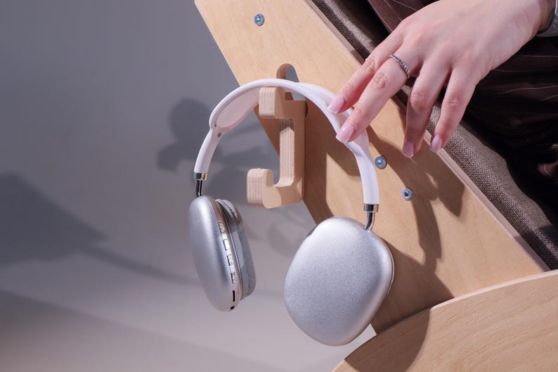 May include: Silver and white over-ear headphones hanging on a wooden headphone stand. The headphones have a white headband and silver earcups. The stand is made of light-colored wood and is mounted on a wooden surface.