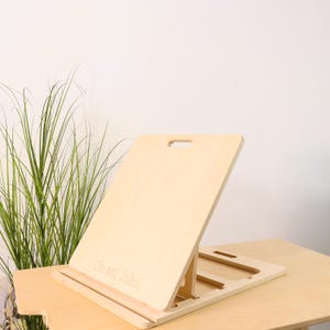 以下が含まれることがあります： 傾斜した描画面を備えた、明るい色の木製イーゼル。 イーゼルは長方形のベースとハンドル付きの背面サポートを備えています。 「Do art, John」の文字が前面に刻まれています。 イーゼルは明るい色の木製のテーブルの上にあります。