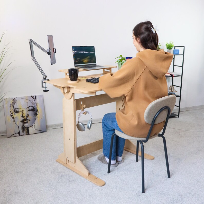 May include: A light-colored wooden standing desk with a laptop, keyboard, and tablet holder. A black mug, headphones, and a black chair are also present. A person is sitting at the desk wearing a brown hooded jacket and blue jeans.