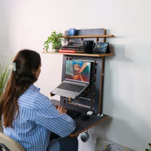 May include: A black and wood wall-mounted desk with a laptop, keyboard, and mouse. The desk has two shelves, one with books and a plant, and the other with a mug and a small calendar. A person is sitting at the desk, using the laptop.