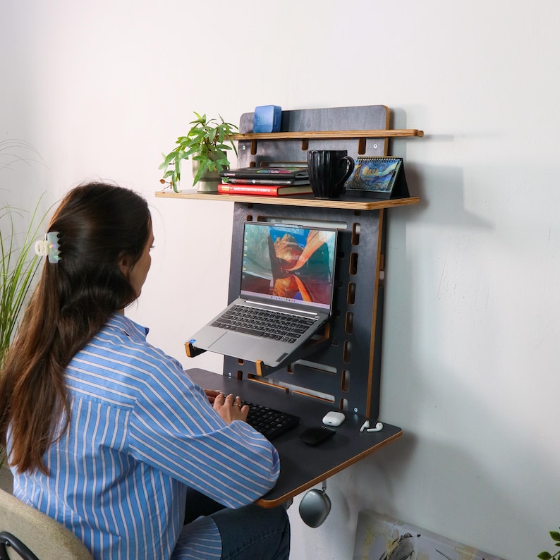 May include: A black and wood wall-mounted desk with a laptop, keyboard, and mouse. The desk has two shelves, one with books and a plant, and the other with a mug and a small calendar. A person is sitting at the desk, using the laptop.