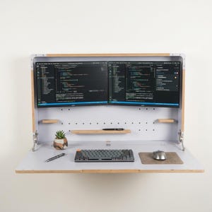May include: A wall-mounted fold-down desk with a light wood frame and white interior. It features two side-by-side computer monitors displaying code, a small potted succulent, a mechanical keyboard, a computer mouse on a pad, and a pen.