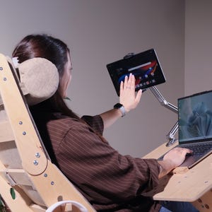 May include: A wooden ergonomic chair with a laptop and tablet stand. The chair has a light wood finish and a beige headrest. A laptop and tablet are mounted on adjustable arms. A smartphone rests on the desk.