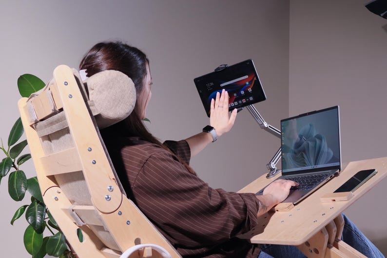 May include: A wooden ergonomic chair with a laptop and tablet stand. The chair has a light wood finish and a beige headrest. A laptop and tablet are mounted on adjustable arms. A smartphone rests on the desk.