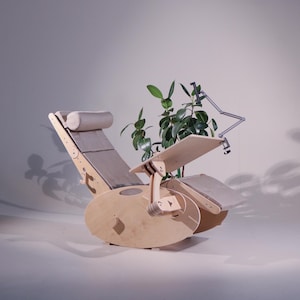K&ouml;nnte beinhalten: Ein h&ouml;lzerner Relaxsessel mit eingebautem Schreibtisch und Fu&szlig;st&uuml;tze. Der Stuhl hat ein hellgraues Kissen und eine Kopfst&uuml;tze. Eine kleine Pflanze steht auf dem Schreibtisch. Ein verstellbarer Metallarm h&auml;lt eine Lampe. Der Stuhl ist aus hellem Holz gefertigt.
