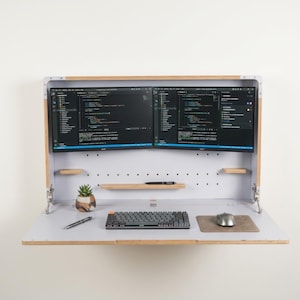 May include: A wall-mounted fold-down desk with a light wood frame and white interior. It features two side-by-side computer monitors displaying code, a small potted succulent, a mechanical keyboard, a computer mouse on a pad, and a pen.