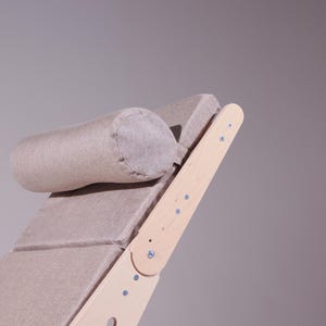 May include: A light beige, adjustable wooden chaise lounge with a matching cylindrical headrest. The frame is made of light-colored wood, and the seat and headrest are upholstered in a textured fabric. The design is minimalist and modern.