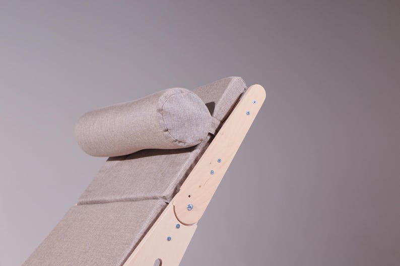 May include: A light beige, adjustable wooden chaise lounge with a matching cylindrical headrest. The frame is made of light-colored wood, and the seat and headrest are upholstered in a textured fabric. The design is minimalist and modern.