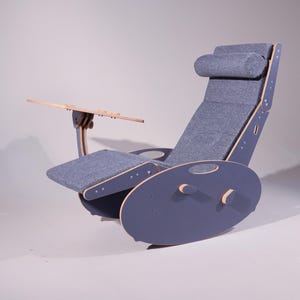 K&ouml;nnte beinhalten: Ein moderner, ergonomischer Schaukelstuhl in einem ged&auml;mpften Blau-Grau. Der Stuhl verf&uuml;gt &uuml;ber eine Kopfst&uuml;tze, eine Armlehne und einen kleinen Holztisch. Sitz und R&uuml;ckenlehne sind mit passendem Stoff bezogen. Die Basis ist zum Schaukeln konzipiert.