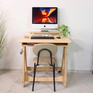 Könnte beinhalten: Ein heller Holzschreibtisch mit Computermonitor, Tastatur und Maus. Vor dem Schreibtisch steht ein kleiner Stuhl mit beigem Sitz und schwarzem Rahmen. Eine kleine Pflanze und Kopfhörer sind ebenfalls zu sehen. Der Computerbildschirm zeigt eine farbenfrohe Landschaft.