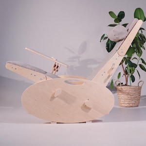 May include: A light-colored wooden rocking chair with a curved base and adjustable backrest. The chair features a gray cushioned seat and headrest, and a small wooden table. A potted plant is in the background.