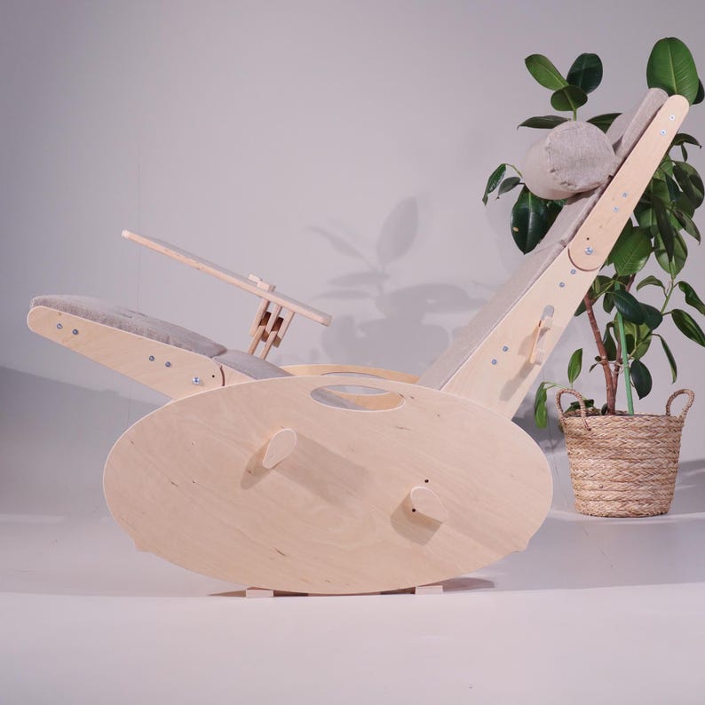 May include: A light-colored wooden rocking chair with a curved base and adjustable backrest. The chair features a gray cushioned seat and headrest, and a small wooden table. A potted plant is in the background.