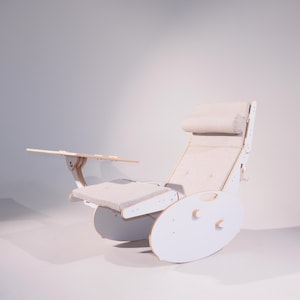May include: A white, modern rocking chair with a built-in side table. The chair features a light gray cushioned seat, back, and headrest. The side table is made of light wood. The chair is designed for comfort and relaxation.