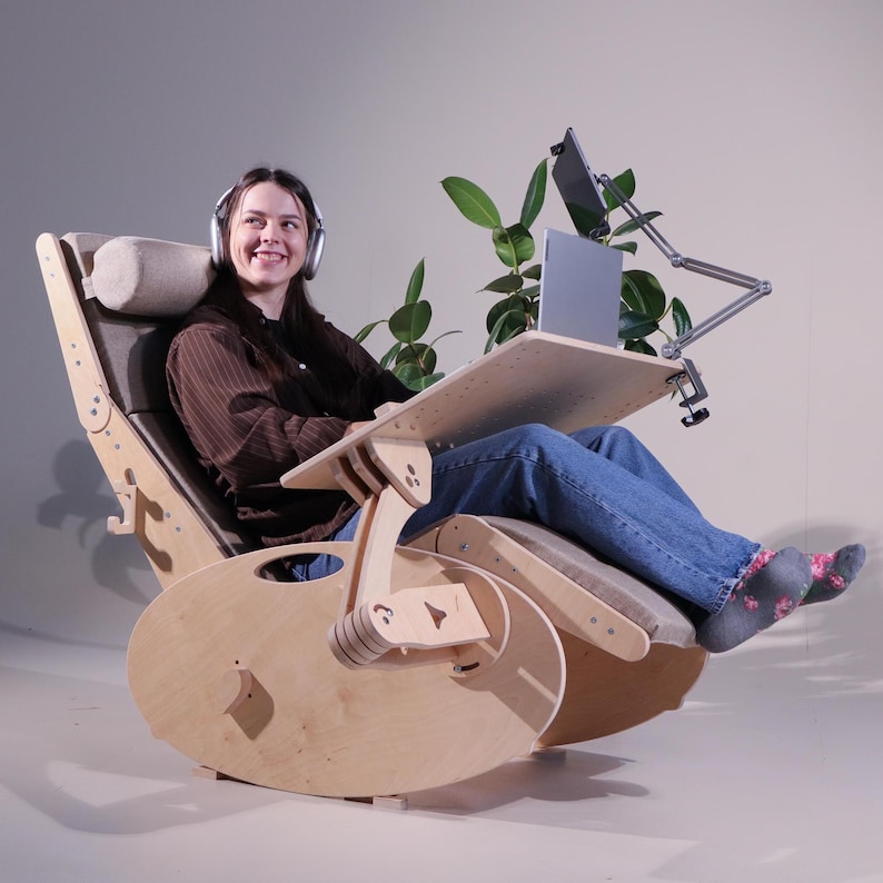 May include: A wooden rocking chair with a built-in desk and adjustable arm for a laptop and tablet. The chair has a light wood finish, gray cushions, and a headrest. The person in the chair is wearing headphones and jeans.