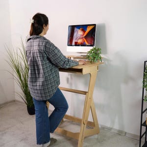 Peut inclure: Un bureau debout en bois clair doté d'un cadre de support incliné et d'une surface de travail plane. Le bureau supporte un écran d'ordinateur affichant une image de paysage colorée. La structure est en contreplaqué avec des veines visibles.
