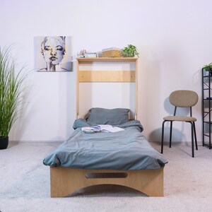 May include: A wooden, single-sized bed with a headboard shelf, featuring a gray duvet and pillow. The bed frame has a curved base. A painting and a chair are visible in the background. The bed is designed for small spaces.